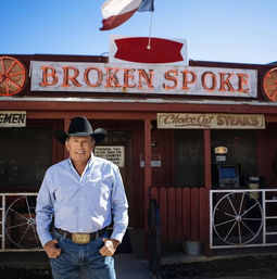 Man in a cowboy hat and belt buckle smiling in front of a rustic Western saloon-style steakhouse storefront with a neon sign, wagon-wheel decor, and a flag against a blue sky.