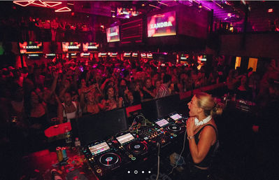Female DJ performing from a crowded DJ booth in a neon-lit nightclub, colorful lights and cheering dancefloor energy
