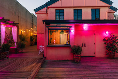 Vibrant evening outdoor patio with a pink neon‑lit building and open service window, wooden deck, string lights, curtained pergola, and potted tropical plants.