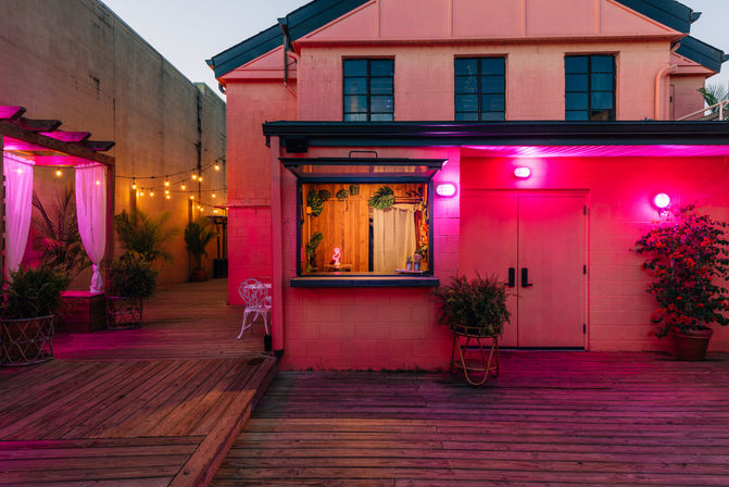 Vibrant evening outdoor patio with a pink neon‑lit building and open service window, wooden deck, string lights, curtained pergola, and potted tropical plants.