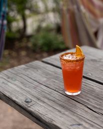 Bright orange iced cocktail in a tall glass with a chili-salt rim and dried orange garnish, sitting on a weathered wooden picnic table on an outdoor patio with blurred greenery