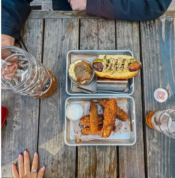 Overhead view of a casual outdoor patio meal on a weathered wooden picnic table: a sausage sandwich with green sauce and slaw in a metal tray, breaded fried chicken strips with a dipping cup in another tray, large beer glasses and diners’ hands visible.