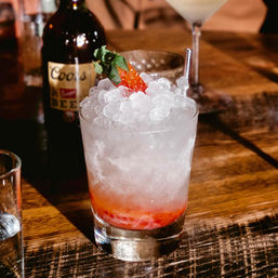 Strawberry crushed-ice cocktail in a rocks glass with metal straw and fresh berry garnish on a wooden bar table, blurred bottle in background.