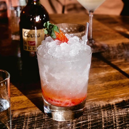Strawberry crushed-ice cocktail in a rocks glass with metal straw and fresh berry garnish on a wooden bar table, blurred bottle in background.