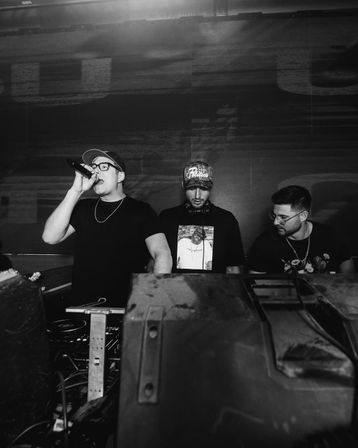 Black-and-white nightclub shot of three DJs behind a booth — left performer rapping into a microphone, center DJ with headphones looking at equipment, right DJ checking gear with a large speaker in the foreground.