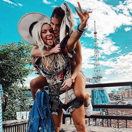Two friends laughing on a sunny urban rooftop — one giving a piggyback and flashing a peace sign with a city skyline and communications tower in the background.
