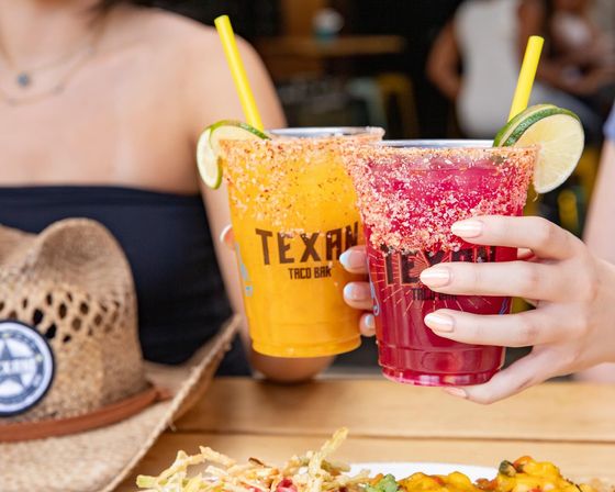 Two colorful frozen cocktails—mango yellow and hibiscus red—in plastic cups with chili-salt rims, lime wedges and yellow straws held over a wooden patio table with Tex‑Mex snacks and a straw cowboy hat nearby.
