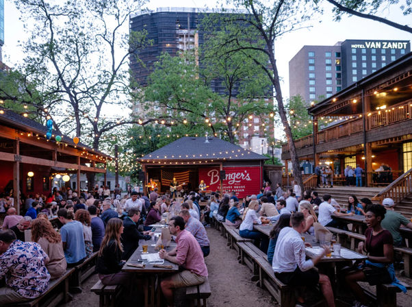 Lively outdoor beer garden at dusk with wooden picnic tables, string lights, a small live-music stage and crowds enjoying drinks beneath city high-rises.