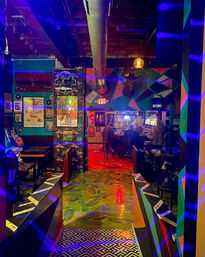 Neon-lit urban bar interior seen from an entryway with bold geometric mural, exposed ductwork, mounted deer head, patterned tile and fern-print floors, framed posters on teal walls, and patrons seated at the back bar.