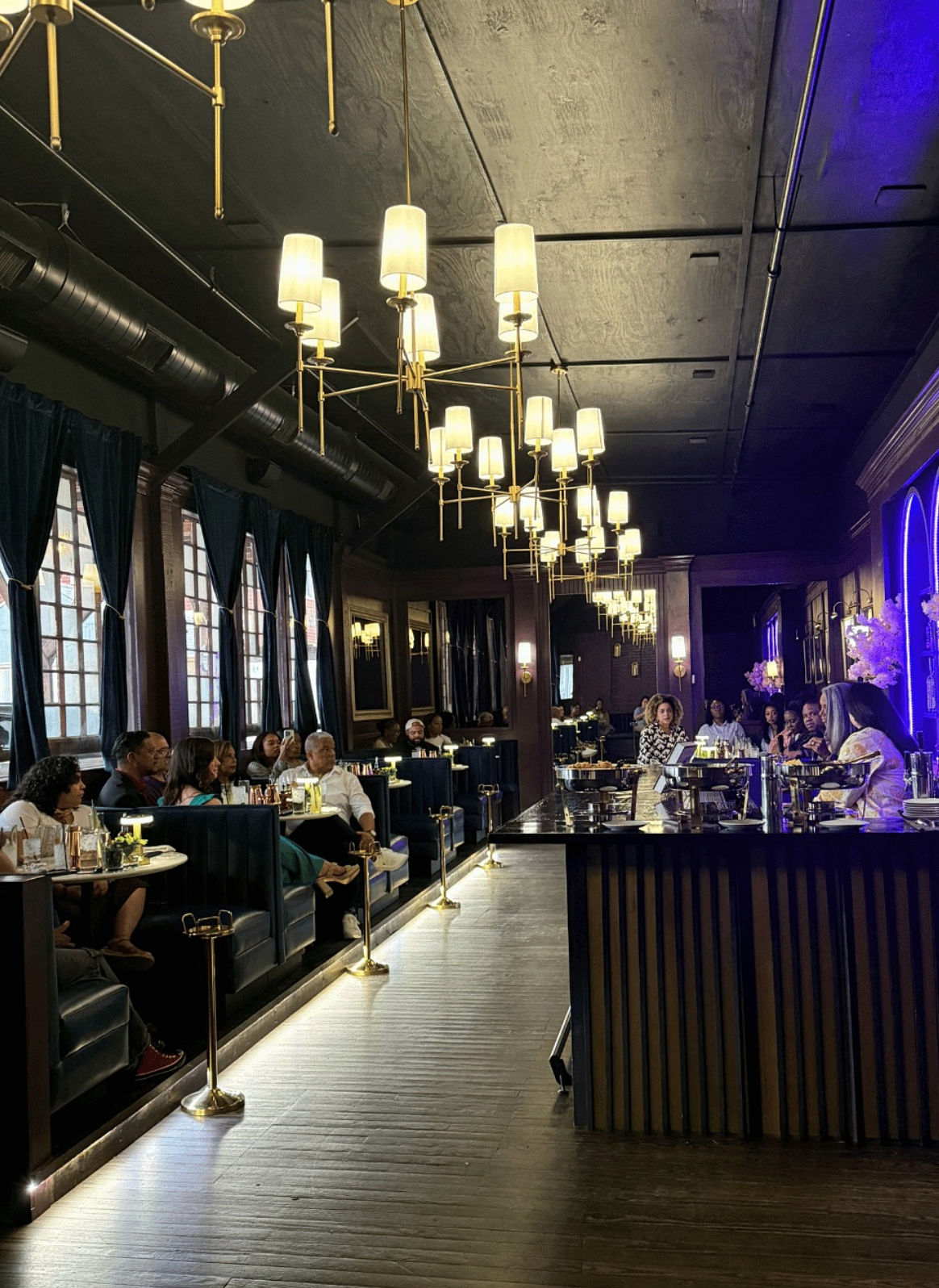 Glamorous urban cocktail lounge interior with brass chandeliers, plush dark booths along tall windows, seated patrons, polished bar counter and purple-accent lighting