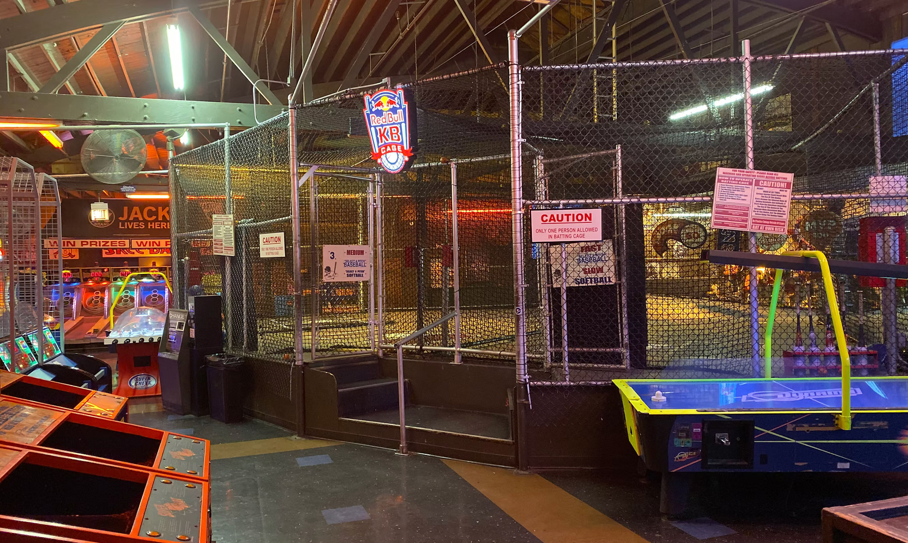 Lively indoor arcade with a fenced batting cage center stage, skee-ball lanes to the left, air hockey table to the right, colorful neon lights and exposed wooden rafters overhead.