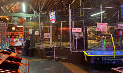 Lively indoor arcade with a fenced batting cage center stage, skee-ball lanes to the left, air hockey table to the right, colorful neon lights and exposed wooden rafters overhead.