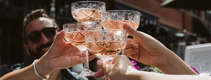 Four cut-crystal coupe glasses of rosé sparkling wine clinking in a sunny outdoor patio toast among friends