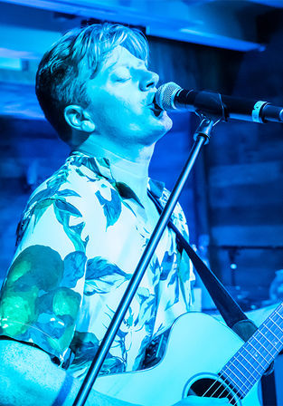 Singer-guitarist performing a live acoustic set on a small indoor stage under blue lighting, wearing a floral shirt and singing into a microphone while playing an acoustic guitar.