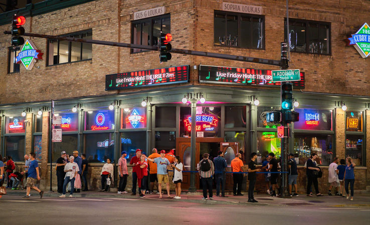 Lively nighttime corner bar on W Addison St in Chicago with brick facade, colorful neon signs, traffic lights, and a crowd waiting outside