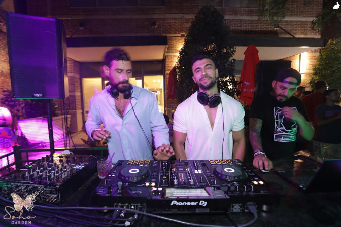 Three DJs performing at an outdoor garden patio party at night, standing behind a professional DJ controller and mixer with colorful purple and green lighting and a lively crowd vibe.