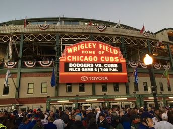 Evening crowd outside historic Wrigley Field in Chicago under a lit marquee reading "Home of Chicago Cubs," announcing Dodgers vs. Cubs NLCS Game 6.