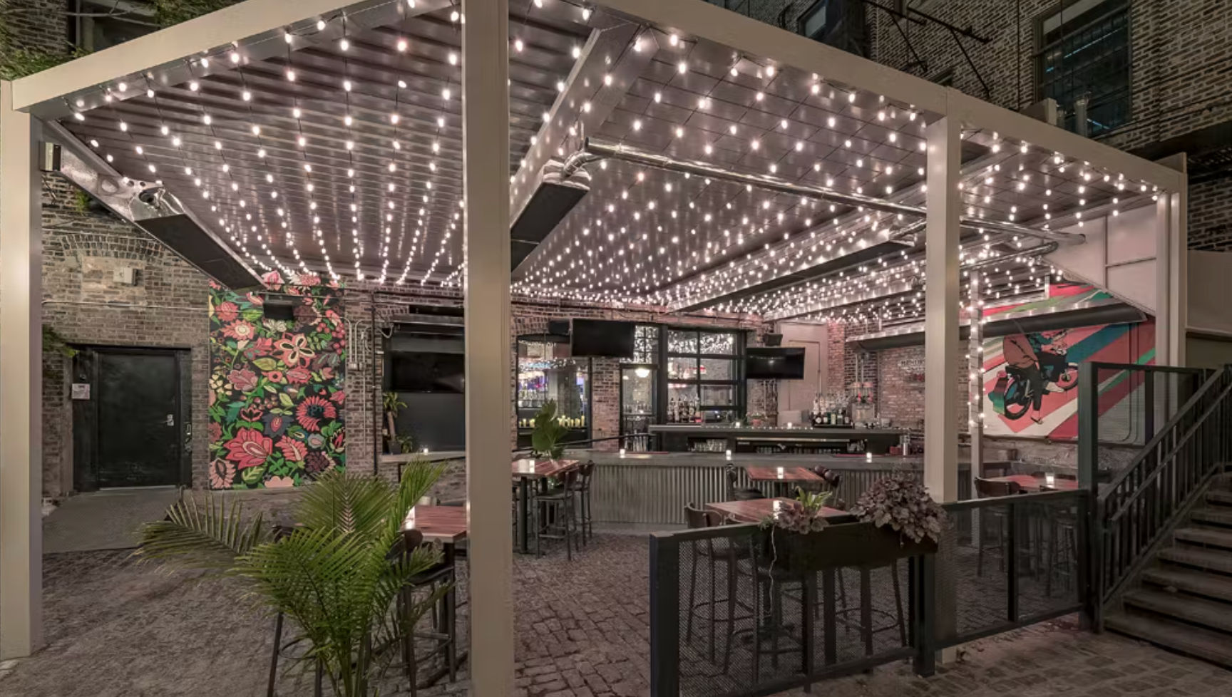 Cozy urban brick courtyard outdoor bar and patio under a pergola strung with warm fairy lights, high-top tables, potted plants, and colorful floral and pop-art murals.