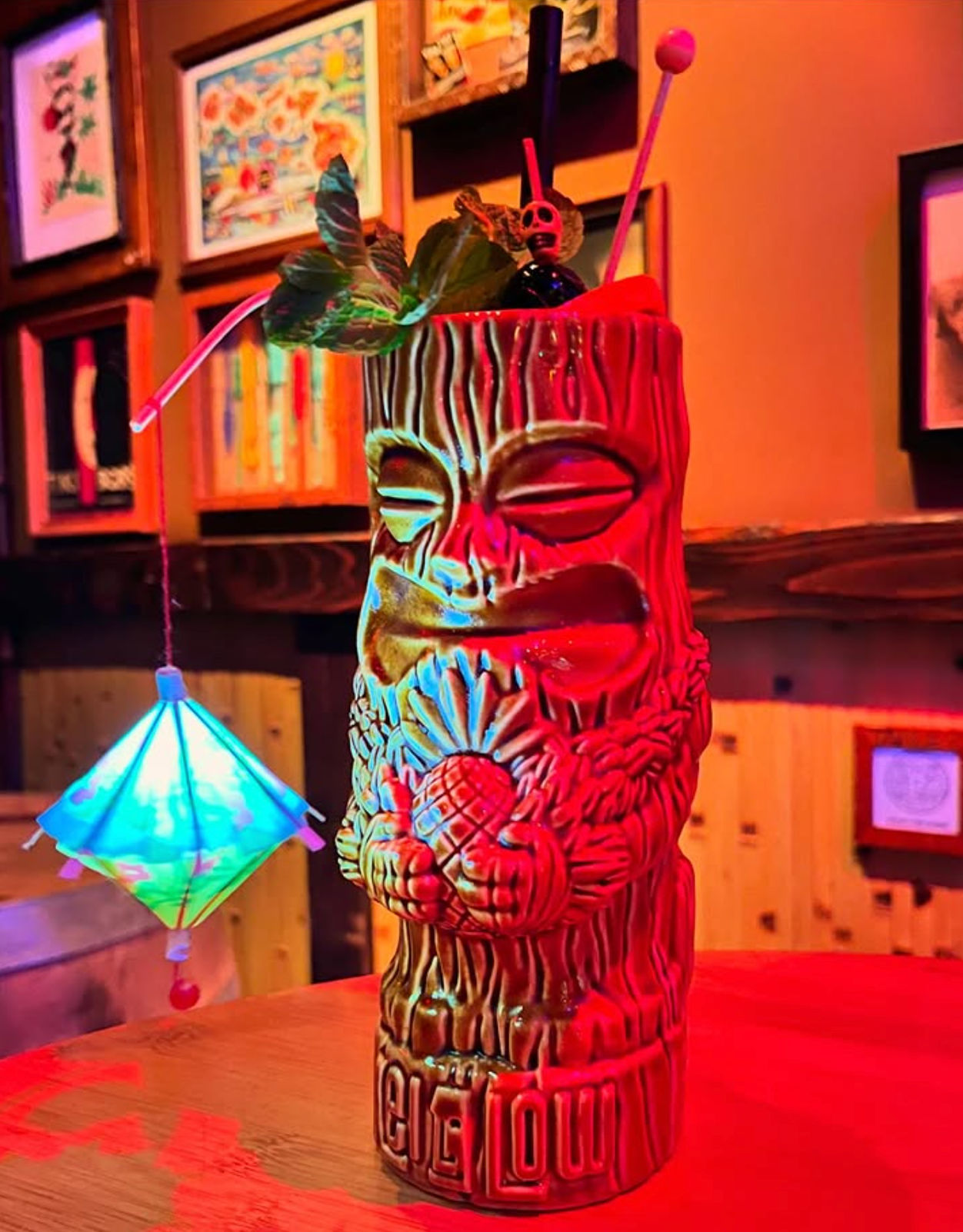 Neon-lit tiki cocktail in a carved ceramic mug with pineapple motif, mint garnish, paper umbrella and swizzle sticks on a wooden bar