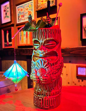 Neon-lit tiki cocktail in a carved ceramic mug with pineapple motif, mint garnish, paper umbrella and swizzle sticks on a wooden bar