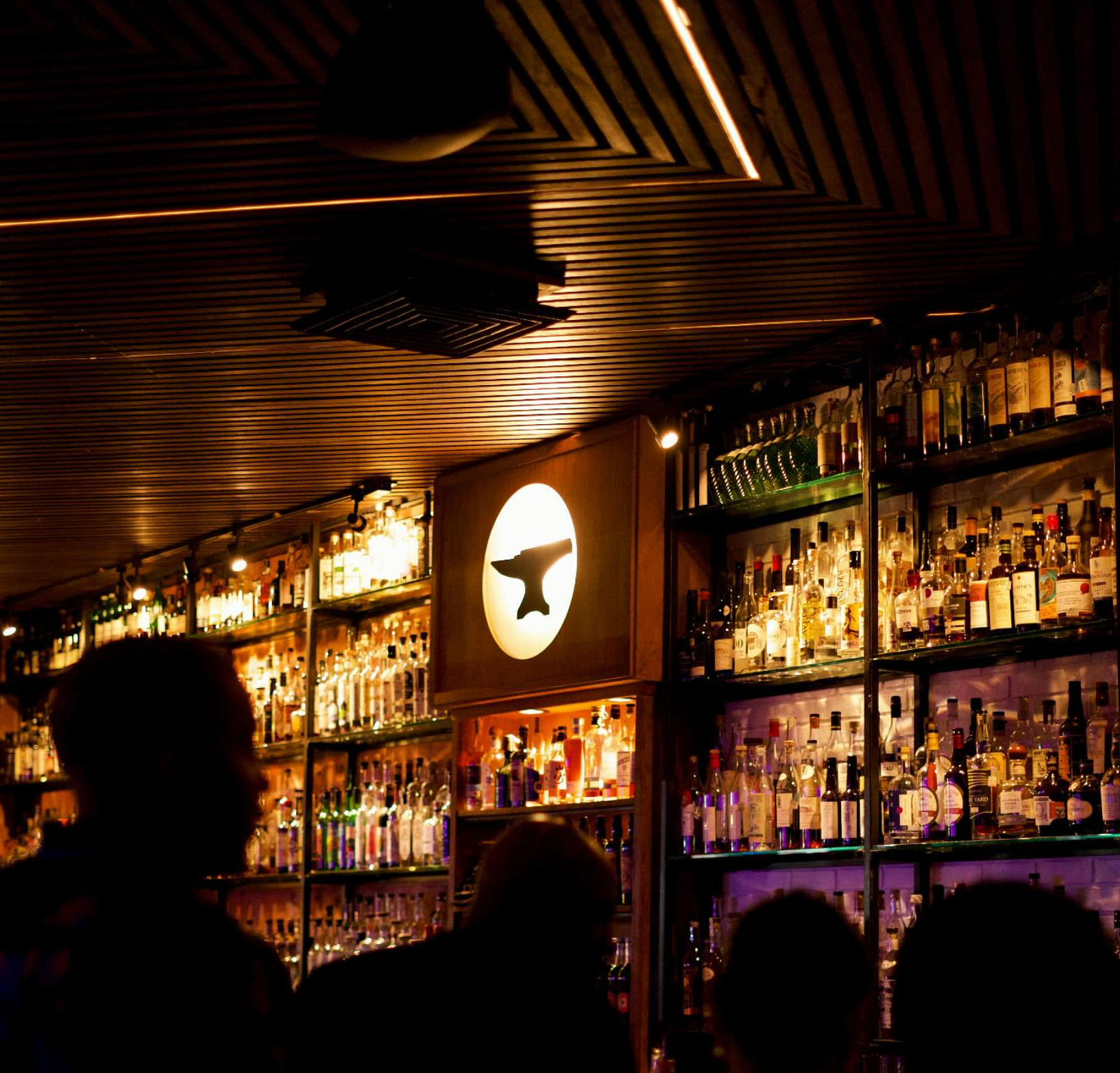Dimly lit cocktail bar interior with backlit shelves of liquor bottles, warm wooden ceiling and silhouetted patrons — cozy urban nightlife scene.