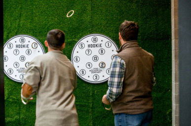 Two players mid-throw doing a ring-toss game at round numbered targets mounted on a green turf wall in an indoor recreation space.