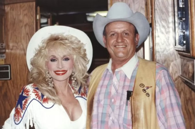 Smiling couple in country-western outfits: woman with big blonde hair in a white fringed dress and cowgirl hat, man in a pastel plaid shirt, tan leather vest and cowboy hat, posed inside a wood-paneled interior.
