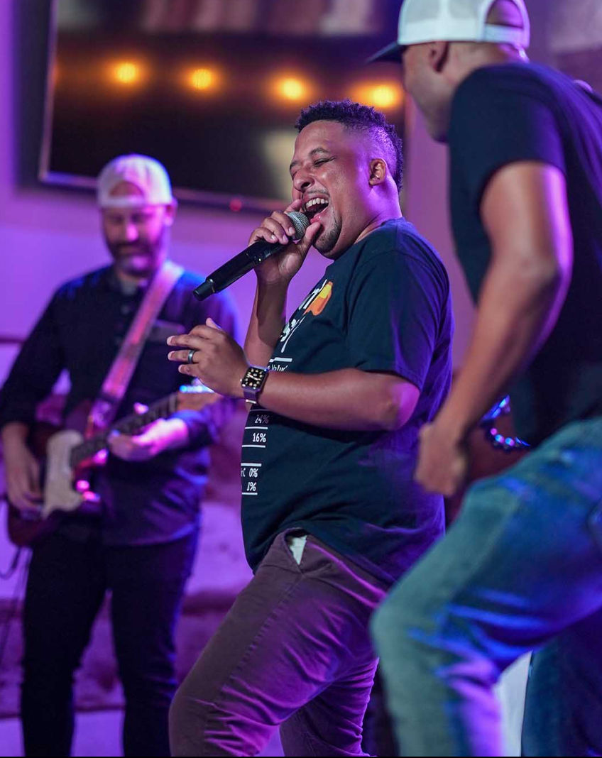 Energetic male singer mid-song holding a microphone on a purple-lit indoor stage, backed by a guitarist and fellow performer at a lively live music venue