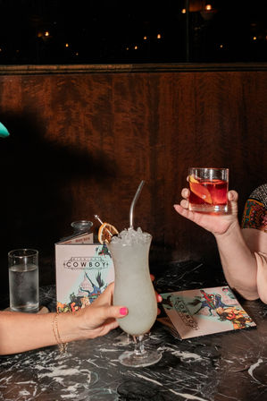 Two hands toasting cocktails at a dim bar — a frosty crushed-ice cocktail in a hurricane glass with metal straw and dried citrus, and an amber drink in a rocks glass with orange peel, on a black marble table with a water glass and colorful illustrated menus.