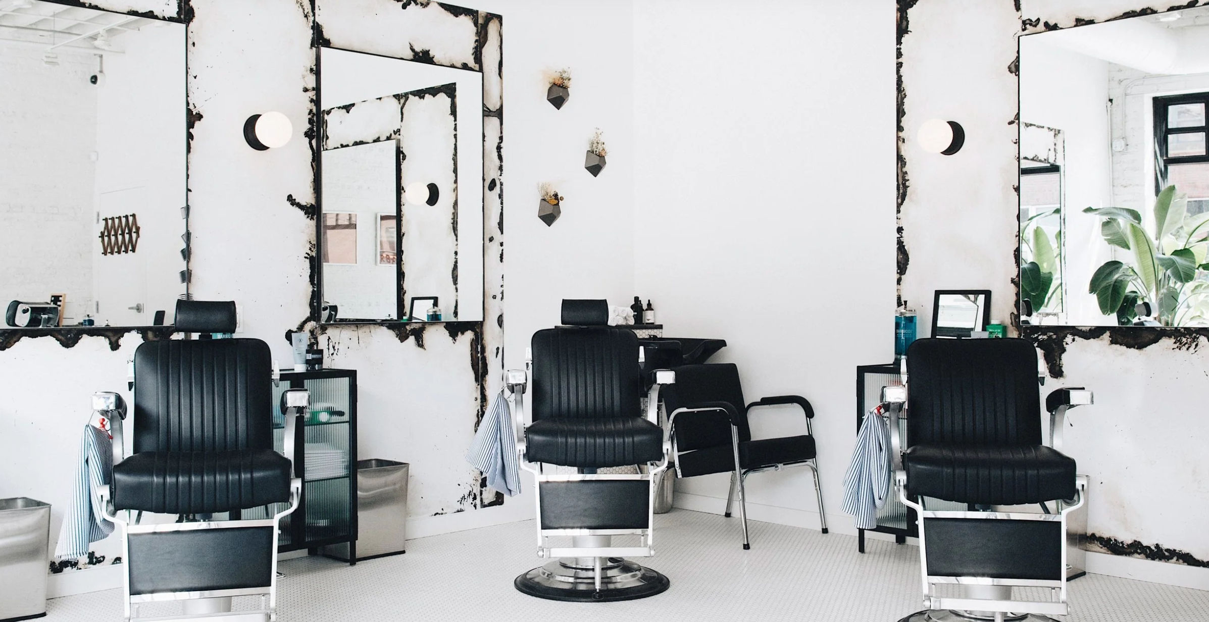 Bright minimalist barbershop interior with three black barber chairs, distressed wall mirrors, white tile floor and leafy plants by the window