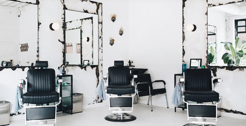 Bright minimalist barbershop interior with three black barber chairs, distressed wall mirrors, white tile floor and leafy plants by the window