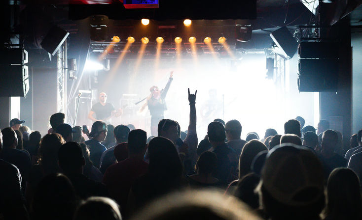 Silhouetted crowd at an indoor concert venue watching a lead singer and band on a brightly backlit stage with golden stage lights and a fan flashing the rock hand