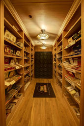 Amber-lit walk-in humidor with floor-to-ceiling wooden shelves stocked with cigar boxes, hardwood floor, decorative ceiling lights and a secured black door at the far end.
