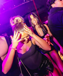 Friends dancing on a crowded nightclub dance floor under purple-pink lights — woman with a topknot holding her head and laughing, another smiling behind her.