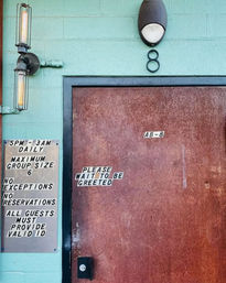 Colorful exterior entrance: teal cinderblock wall with a rust-red metal door numbered 8, retro industrial wall sconce, and signs reading “Please wait to be greeted” plus posted hours and rules (5PM–3AM, max group size 6, valid ID required).