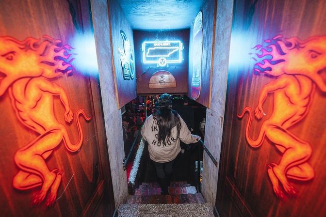 Neon-lit basement bar entrance at night, a person descending narrow stairs toward a crowded dance floor flanked by glowing orange flame-creature murals and bright neon signs