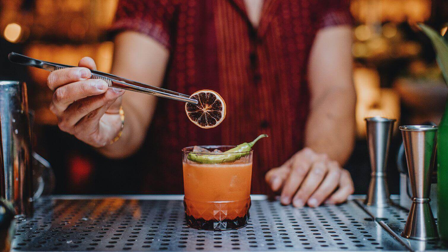 Bartender using metal tongs to place a dried citrus wheel on a bright orange craft cocktail topped with a whole green chili, on a perforated metal bar counter