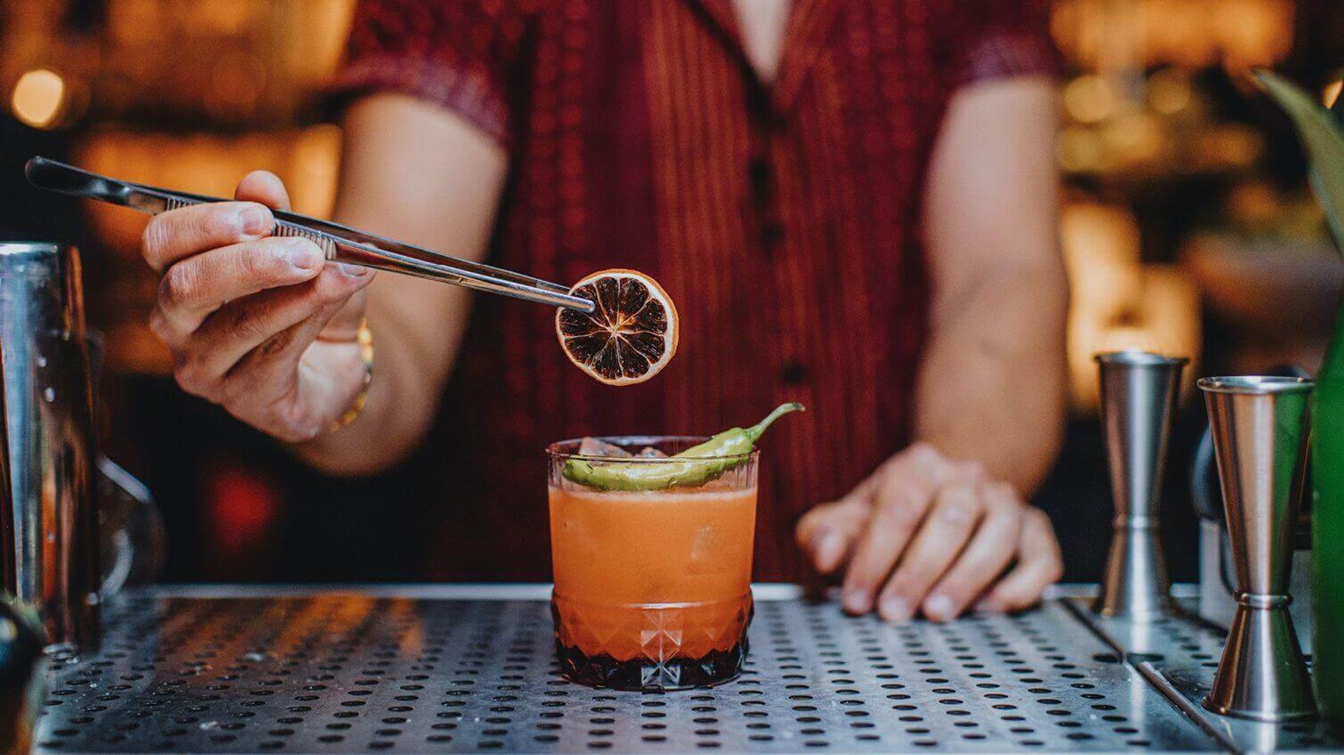 Bartender using metal tongs to place a dried citrus wheel on a bright orange craft cocktail topped with a whole green chili, on a perforated metal bar counter