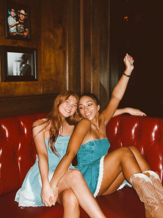 Two smiling friends in blue denim mini dresses sitting close on a red leather booth in a wood-paneled lounge, one wearing cowboy boots with her arm raised, enjoying a night out.
