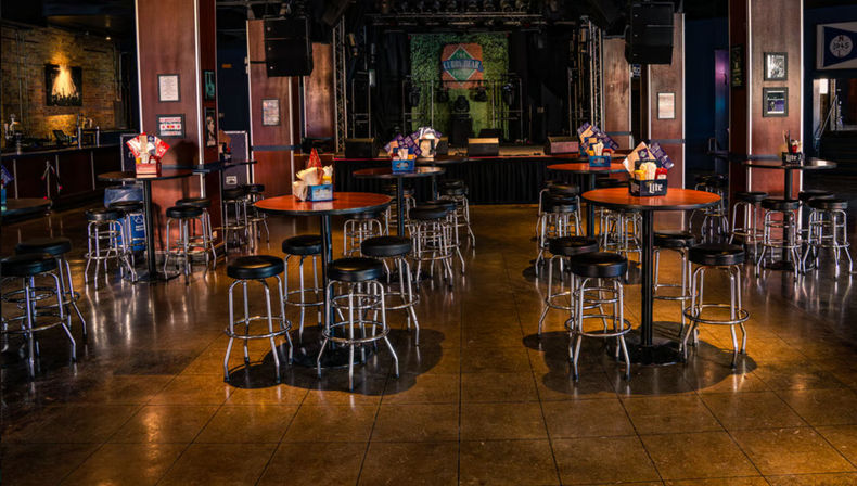 Empty indoor live music venue with round wooden high-top tables and metal bar stools on a tiled floor facing a small stage with speakers and a colorful backdrop, dimly lit and ready for a show.