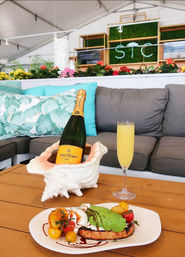 Champagne bottle chilling in a shell-shaped holder with a mimosa glass and avocado toast with cherry tomatoes and burrata on a wooden table in a bright outdoor lounge with tropical pillows and flowering planters — brunch scene