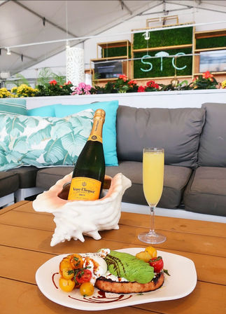 Champagne bottle chilling in a shell-shaped holder with a mimosa glass and avocado toast with cherry tomatoes and burrata on a wooden table in a bright outdoor lounge with tropical pillows and flowering planters — brunch scene