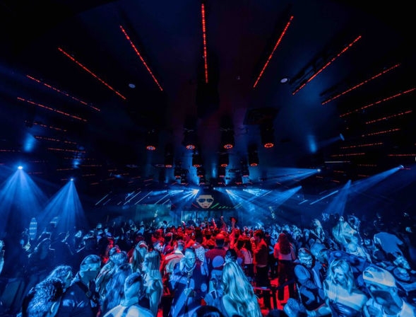 Packed nightclub dance floor with a lively crowd dancing under dramatic blue and red stage lights, laser beams and ceiling LED strips, DJ visuals on stage.
