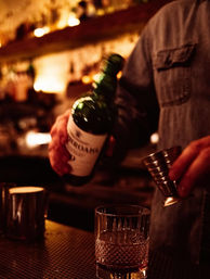 Bartender in a cozy dimly lit cocktail bar pouring whiskey from a green bottle into a metal jigger above a cut-glass rocks glass, warm bokeh shelves in the background.