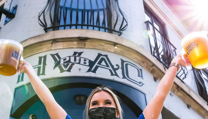 Masked person raising two pints of beer outside a sunny downtown pub entrance with white brick facade and decorative iron balcony