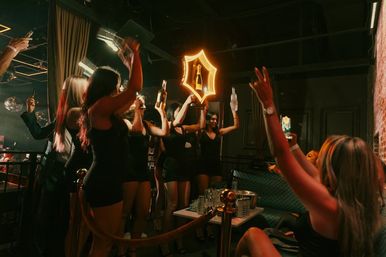 Women in black dresses at a nightclub VIP table raising bottles in celebration under a glowing neon bottle sign, with dim lounge lighting and disco balls