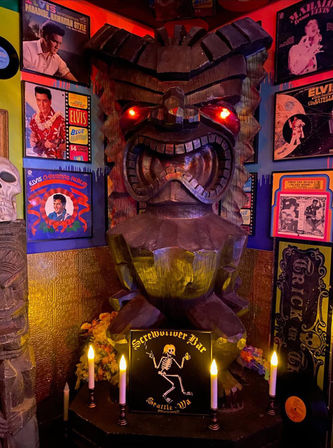 Carved wooden tiki statue with glowing red eyes in a dim retro bar corner, surrounded by vintage vinyl album covers, candles, skull decor and a dancing skeleton sign