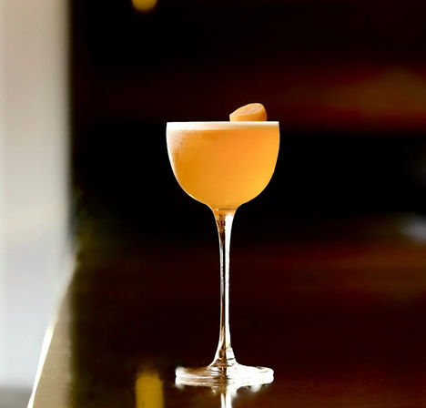 Golden frothy craft cocktail in an elegant stemmed coupe, topped with an orange peel, on a glossy bar with a moody dark background.