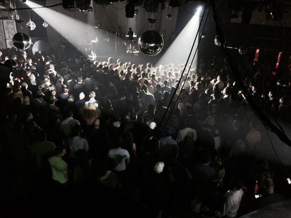 Packed dancefloor in a dim indoor music venue, disco balls glinting as spotlights cut through the crowd toward the stage