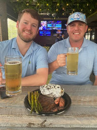 Two smiling men in casual shirts holding large beer mugs at a rustic bar patio, with a plate of grilled steak, asparagus and a foil-wrapped baked potato on a wooden table, TV screens and hanging greenery in the background